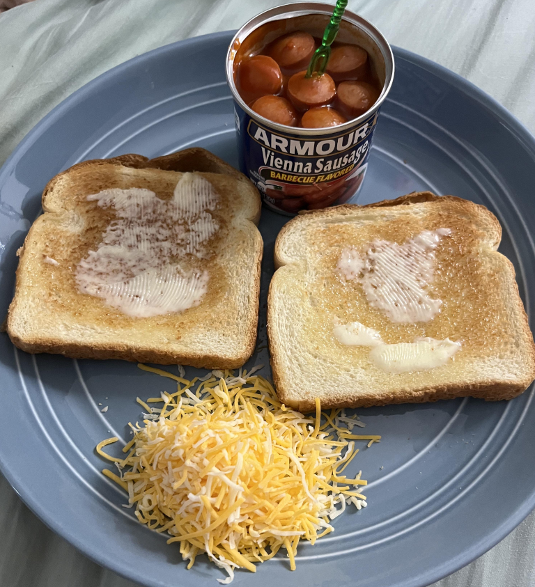 A blue plate with two slices of buttered toast, a small pile of shredded cheese, and an open can of Armour Vienna Sausages in barbecue sauce—classic foods that boomers still eat—served with a green fork in the can.