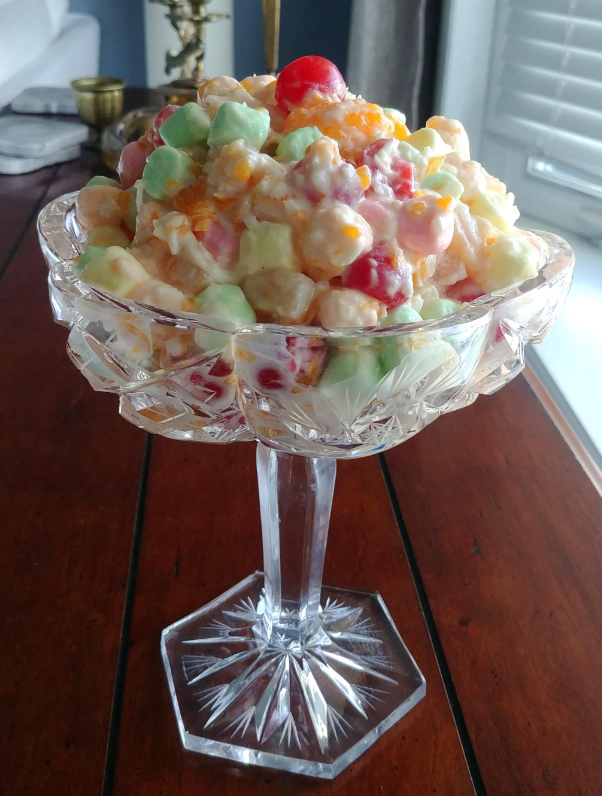 A glass dessert dish filled with colorful ambrosia salad—a classic among foods that boomers still eat—features mini marshmallows, cherries, orange segments, and pineapple chunks in a creamy dressing, sitting on a wooden table near a window.