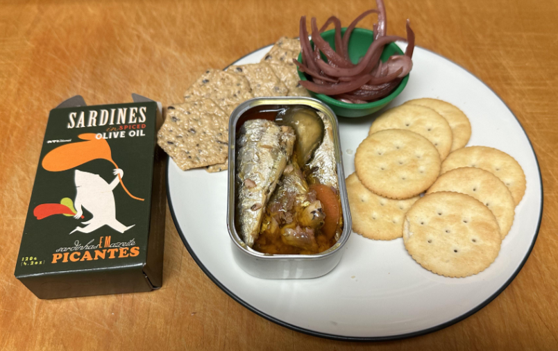 A plate with round crackers, seeded crackers, pickled red onions, and an open tin of sardines in oil—classic foods that boomers still eat—sits next to a box labeled "Sardines in Spiced Olive Oil Picantes" on a wooden surface.