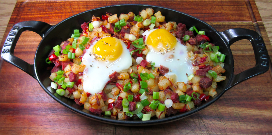 Oval cast iron dish filled with diced potatoes, corned beef, chopped green onions, and red peppers—classic foods that boomers still eat—topped with two sunny-side-up eggs, placed on a wooden cutting board.
