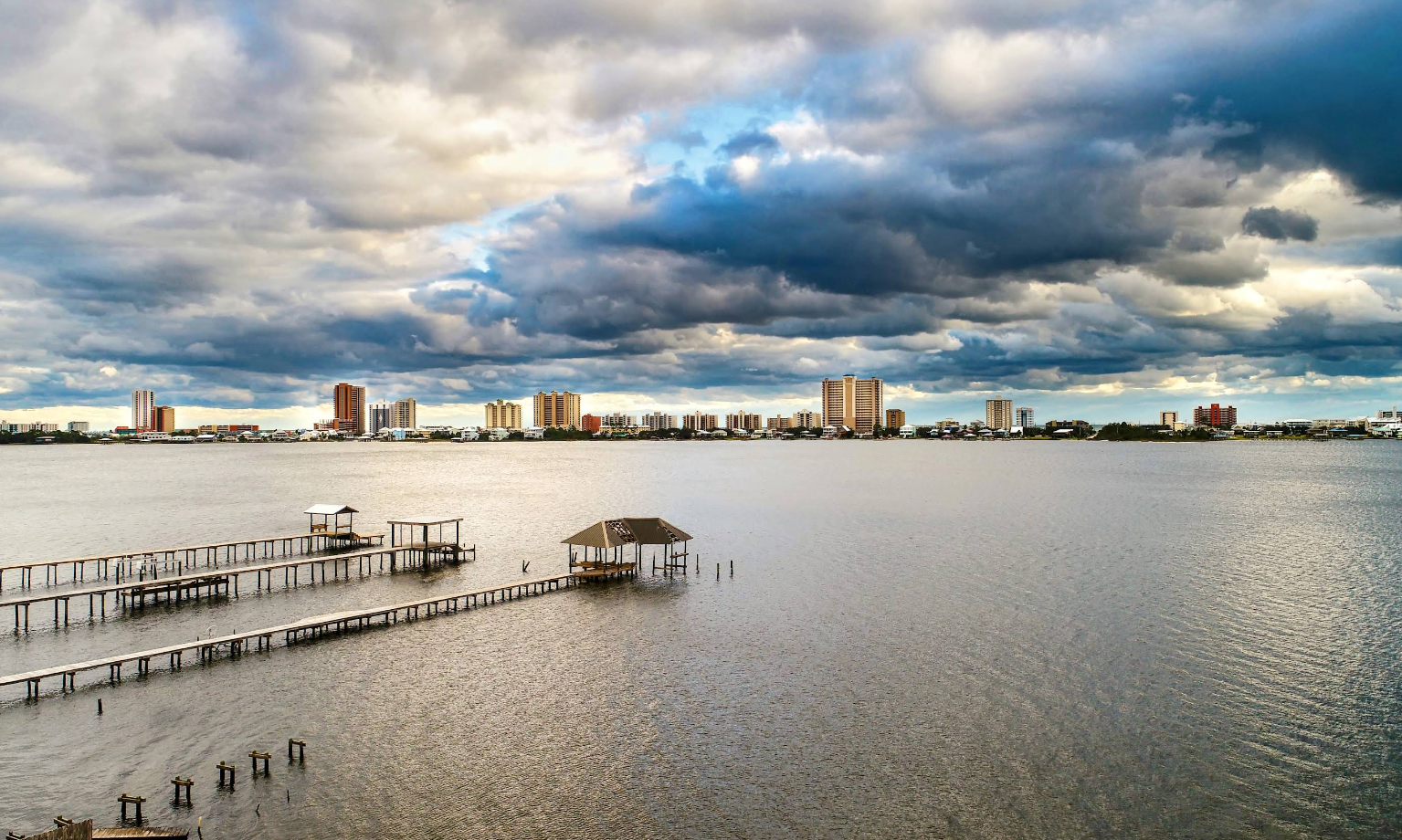 A large body of water with wooden docks and gazebos in the foreground evokes the charm of beach towns, while a distant city skyline and dramatic clouds fill the sky above.