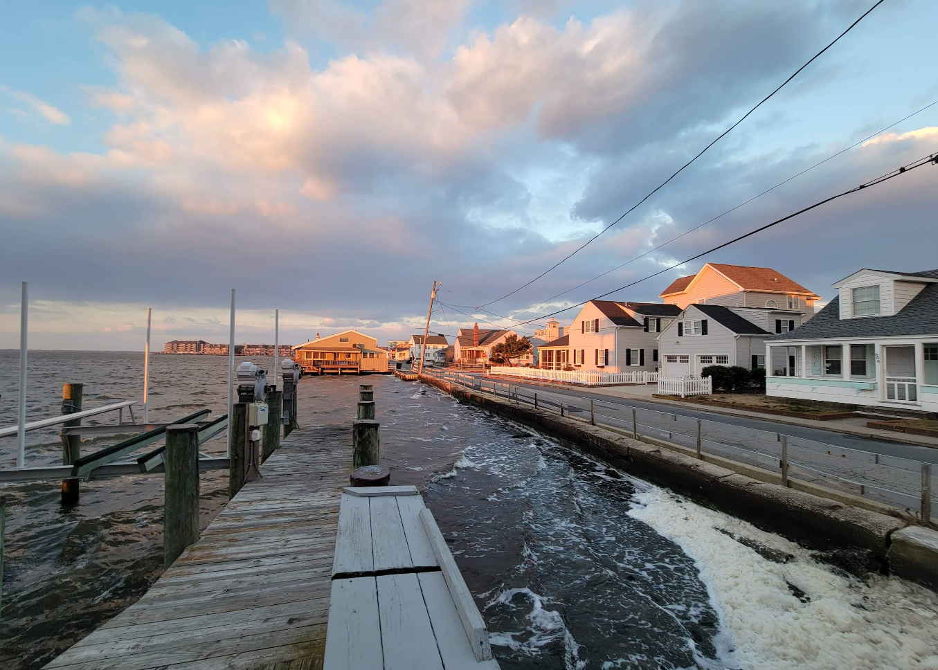 A coastal street in one of the beach towns is flooded, with water covering the road and part of a wooden dock. The sky is partly cloudy, and sunlight illuminates the buildings on the right.