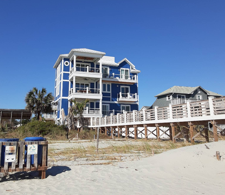 A bright blue, three-story beach house with white trim stands near a sandy beach—capturing the laid-back charm of classic beach towns. A wooden boardwalk leads to the house, with palm trees and grasses under a clear blue sky.