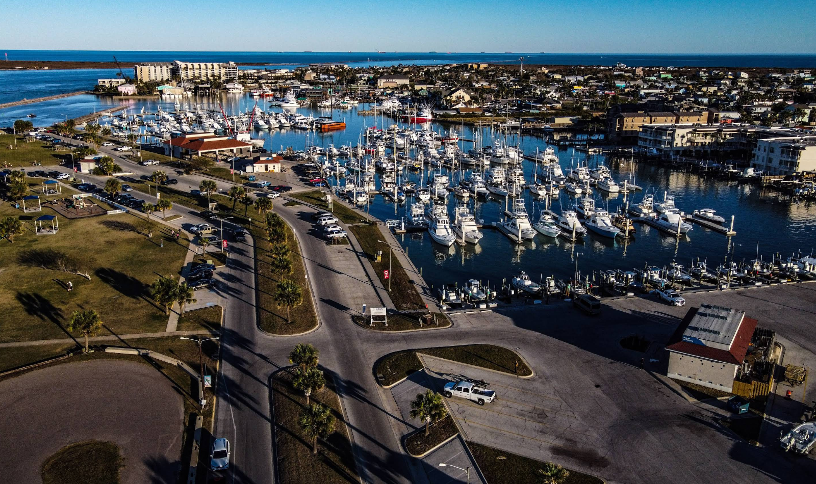 Aerial view of a marina filled with numerous white boats and yachts, surrounded by buildings and roads, with green spaces and palm trees—capturing the relaxed charm of beach towns—with the ocean visible in the distance under a clear sky.