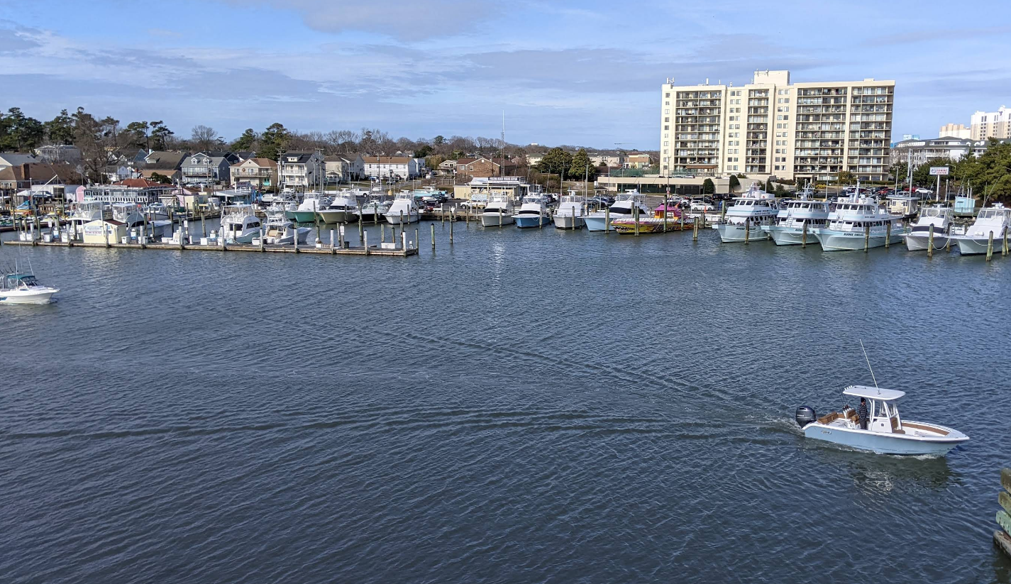 A small motorboat moves across a marina, leaving a wake in the water. Numerous parked boats and yachts are docked along the harbor, evoking the charm of beach towns, with apartment buildings and houses visible under a partly cloudy sky.