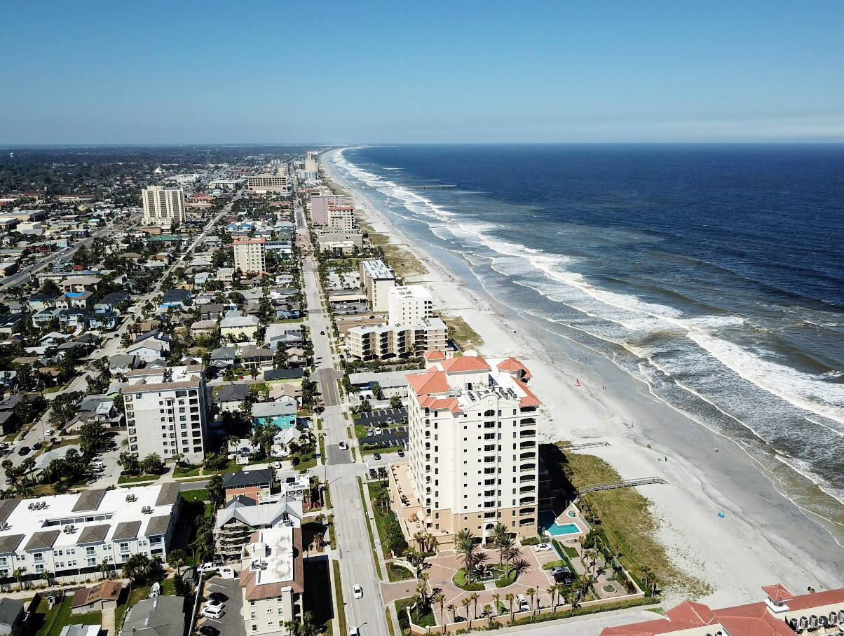 Aerial view of vibrant beach towns with tall buildings lining a sandy shore, waves rolling in beneath a clear blue sky. The cityscape stretches along the coastline in the distance.