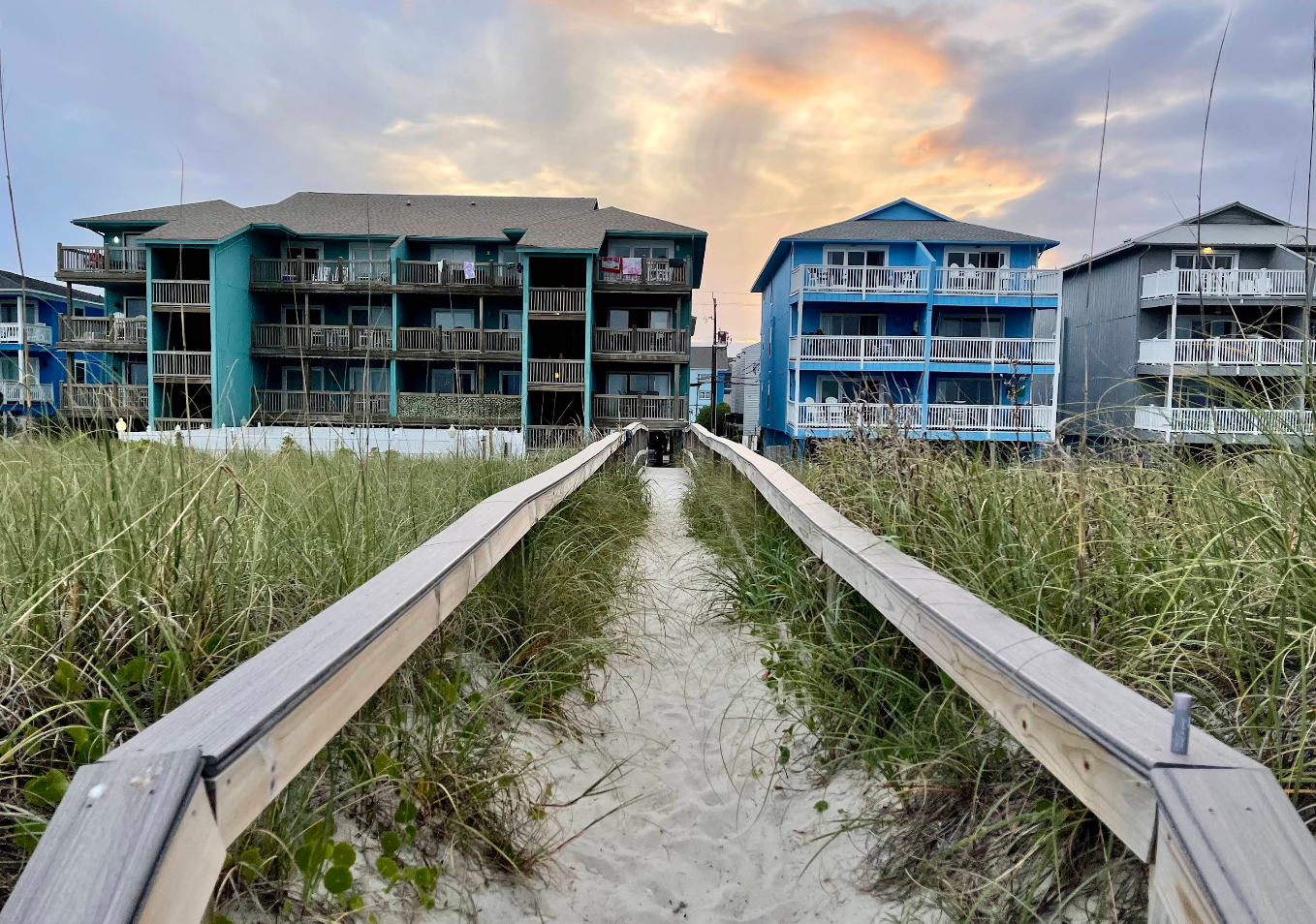 A sandy boardwalk lined with grass leads to coastal condos, capturing the charm of beach towns, with colorful buildings under a dramatic, cloudy sunset sky.