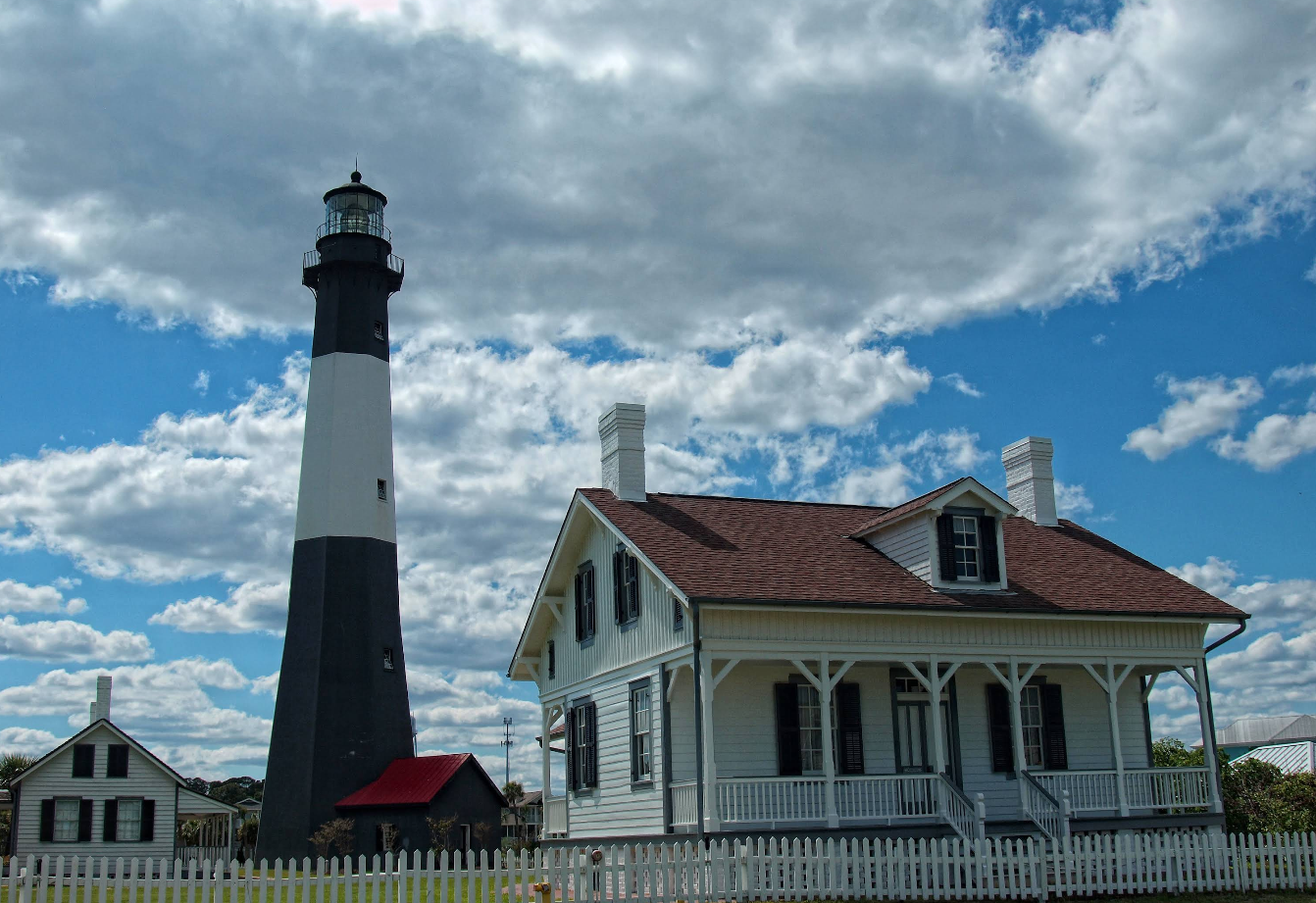 A tall black and white lighthouse stands next to a white house with a red roof and wraparound porch, surrounded by a white picket fence—an iconic sight found in many charming beach towns under a partly cloudy blue sky.