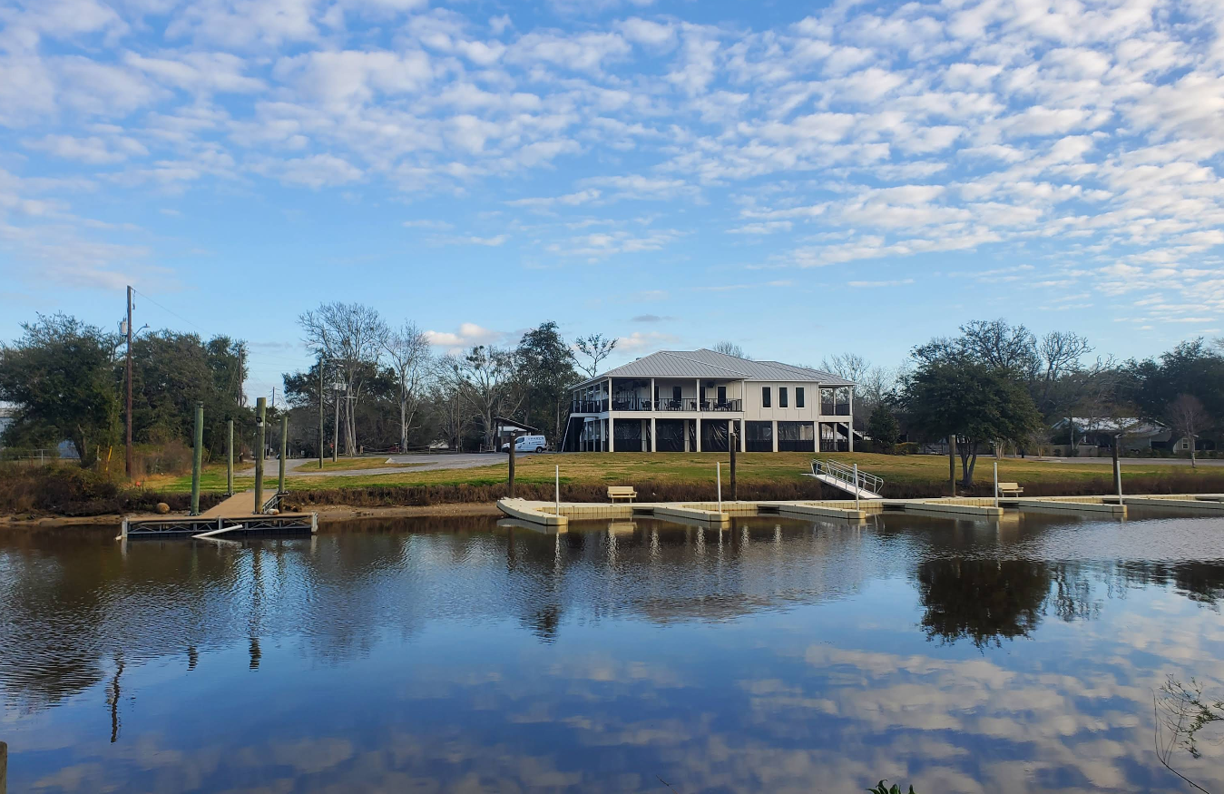 A waterfront building with white walls and a gray roof stands behind a dock and calm water, reflecting the sky with scattered clouds—an inviting scene often found in charming beach towns. Benches and trees line the shore.