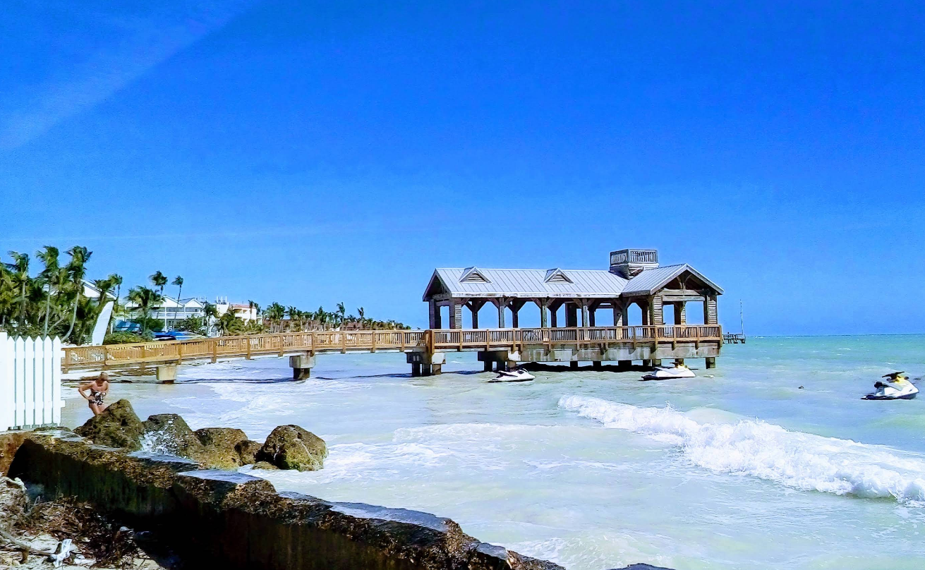 A wooden pier with a covered gazebo extends over turquoise ocean waves, with jet skis nearby. Palm trees and buildings from vibrant beach towns line the distant sandy shore under a clear blue sky.
