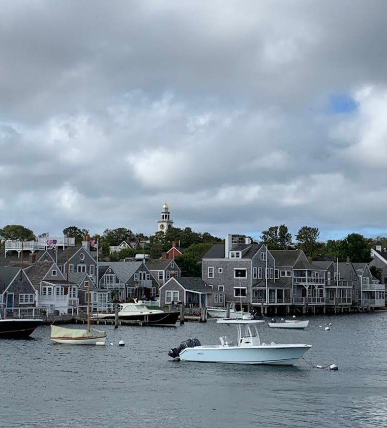 A small white boat floats in a harbor of classic beach towns, with several gray shingled houses and other boats along the shore. A church steeple rises in the background under a cloudy sky.