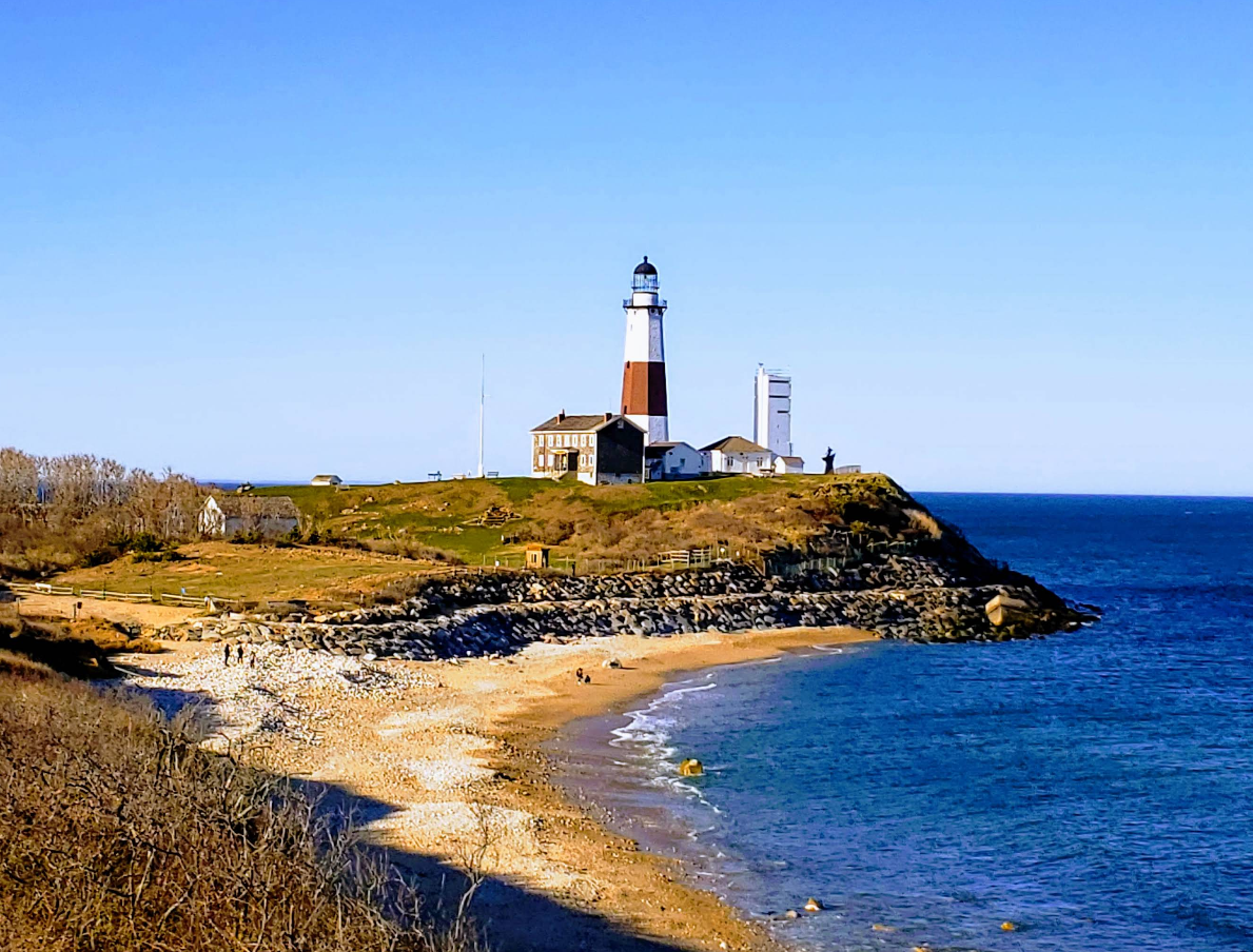 A lighthouse stands on a rocky bluff overlooking the ocean, with sandy beach and blue water in the foreground and several buildings nearby, capturing the charm of classic beach towns under a clear blue sky.