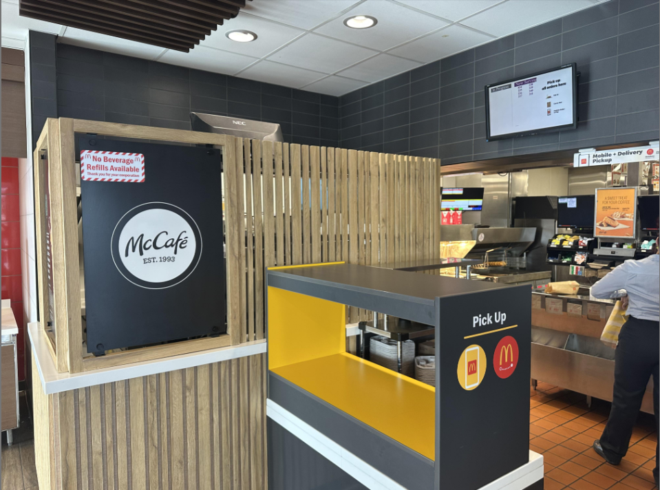 Interior of a McDonald's restaurant showing a McCafé section, a pickup counter with yellow accents, menu screens, and part of the kitchen area with an employee in the background.