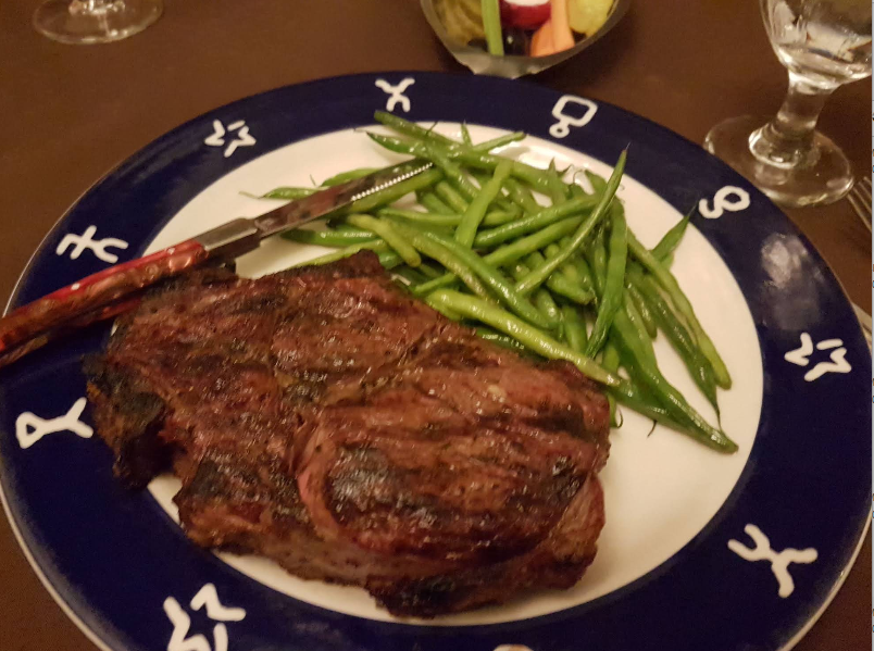 A grilled steak and green beans served on a blue and white plate, with a steak knife on the side—classic fare you might find at a steakhouse on the West Coast. A glass of water and another dish appear partially in the background.