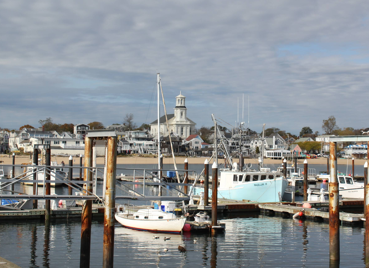 A small harbor with several boats docked at wooden piers, set against classic beach towns scenery. A prominent white church with steeple rises among cozy houses under a cloudy sky.