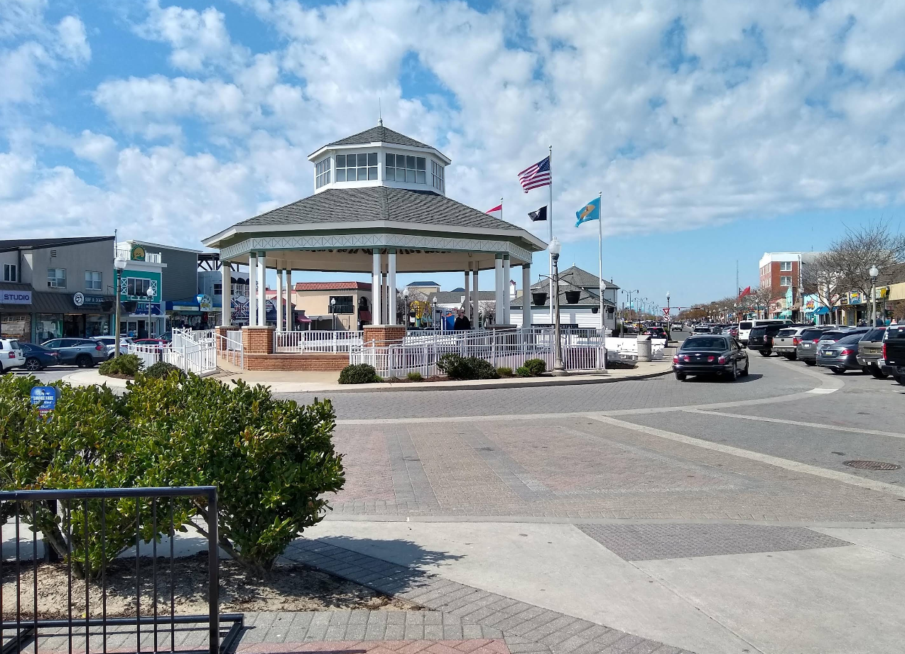 A white gazebo stands in the center of a roundabout, surrounded by shops, parked cars, and flags under a blue sky with scattered clouds—a classic scene found in charming beach towns. Green shrubs are in the foreground.