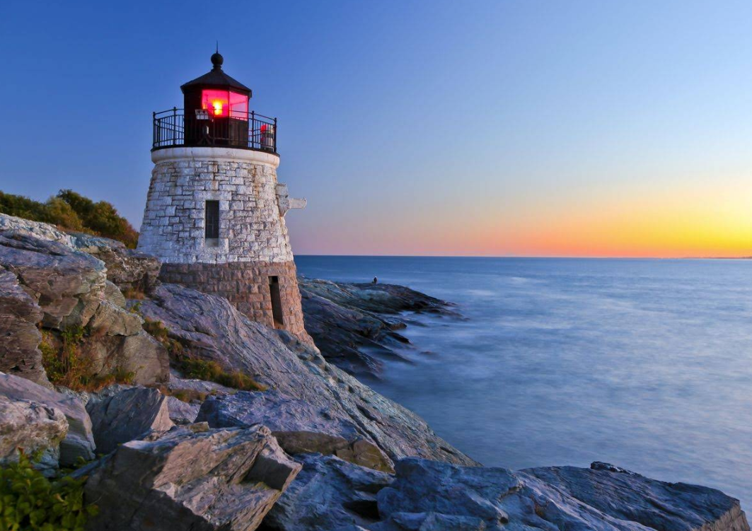 Stone lighthouse with a red light stands on a rocky shore at sunset, overlooking calm blue water and a golden horizon—perfect for exploring nearby beach towns.
