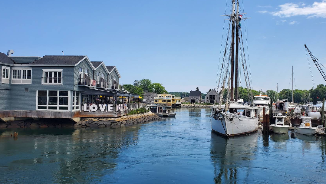 A scenic harbor in one of the charming beach towns, with a sailboat docked near wooden buildings, one displaying large "LOVE" letters. The sky is clear and blue, and smaller boats are moored along the water’s edge.