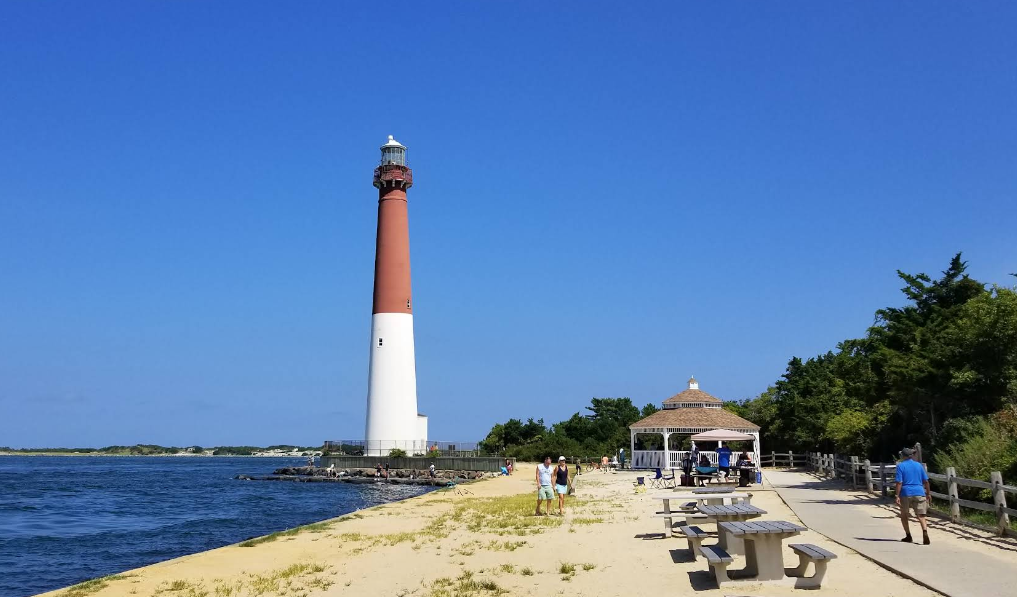 A tall red and white lighthouse stands by the water’s edge on a sandy shore, capturing the charm of beach towns, with people walking, benches, a gazebo, and green trees under a clear blue sky.