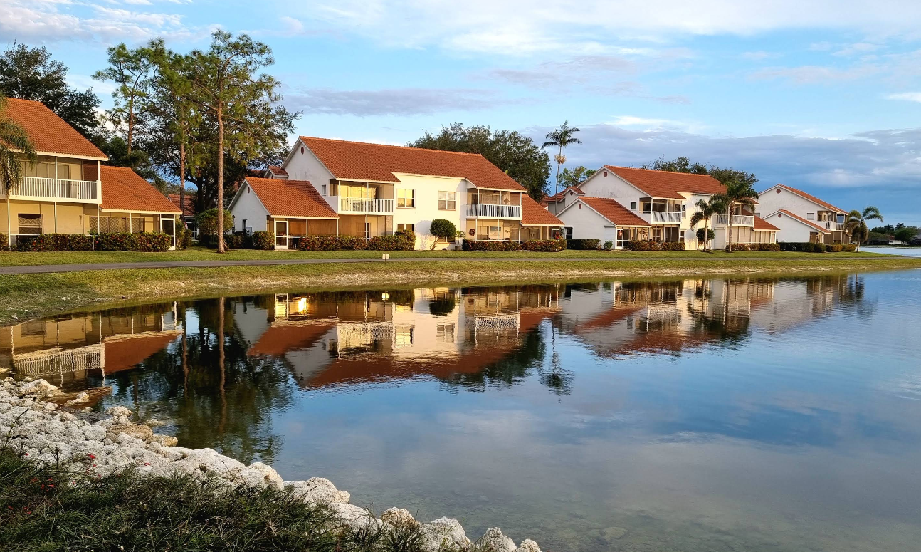 Several houses with red-tiled roofs are reflected in a calm pond, evoking the charm of beach towns, with trees and shrubs along the water’s edge under a partly cloudy sky. Rocks line the shore in the foreground.