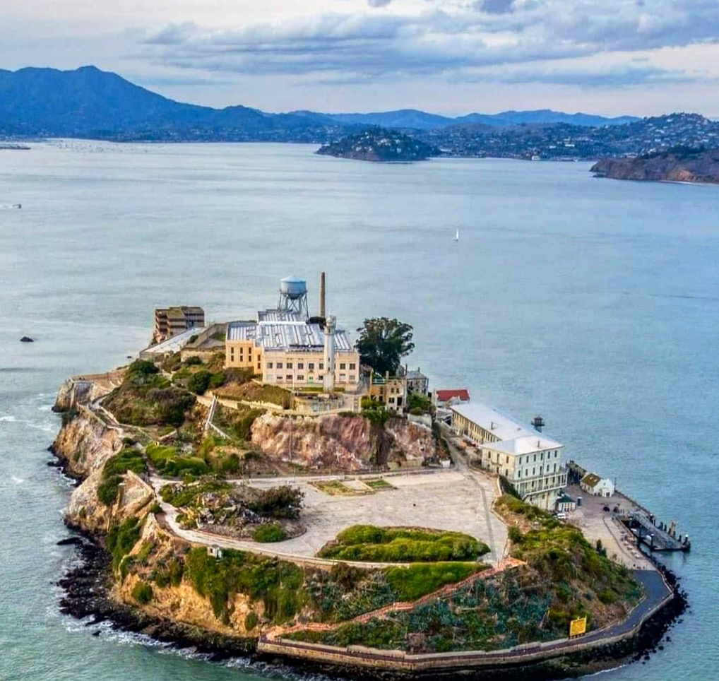 Aerial view of Alcatraz Island in San Francisco Bay, one of the city's top tourist attractions, featuring historic prison buildings surrounded by water, with distant hills and the city visible under a cloudy sky.
