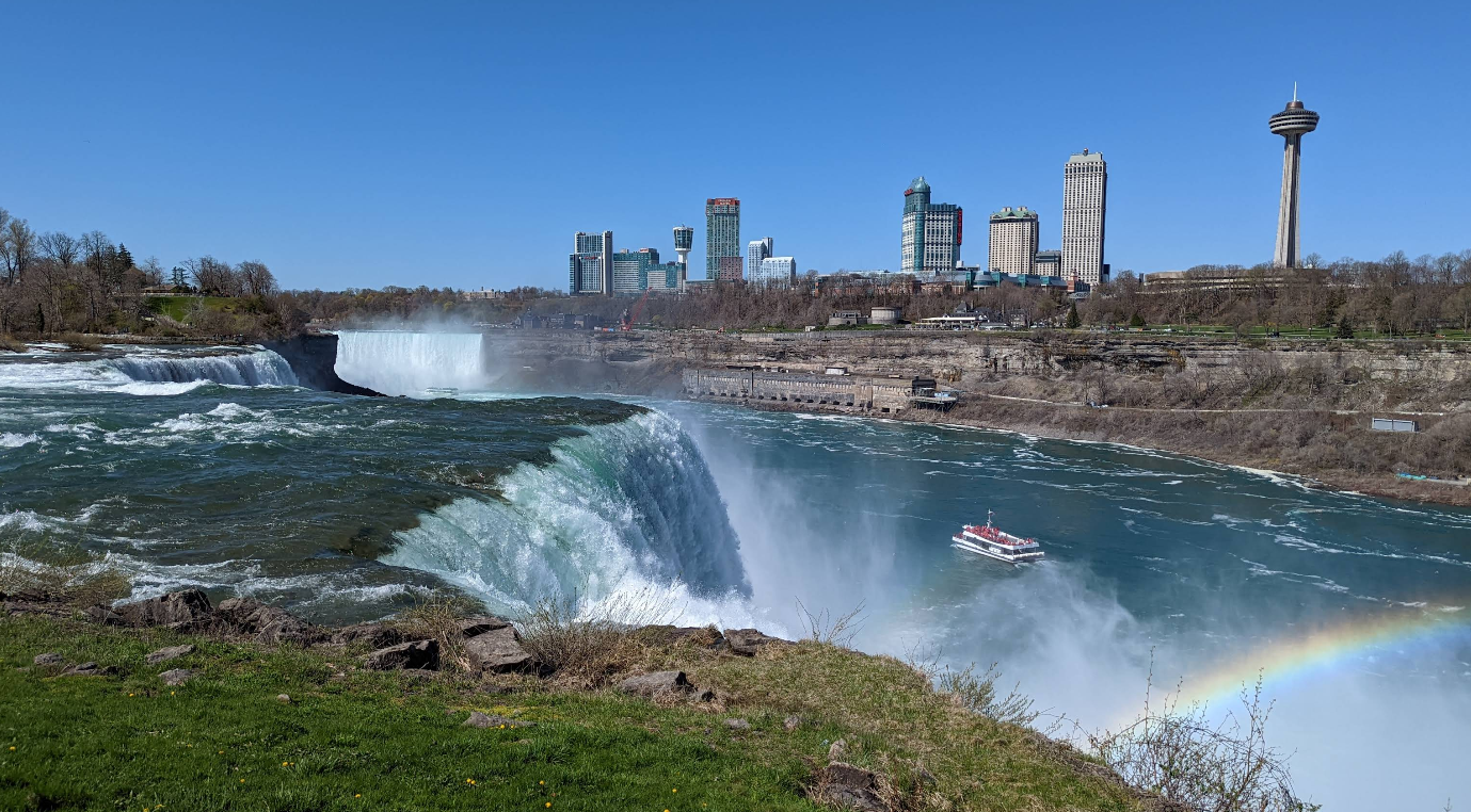 View of Niagara Falls with cascading water, mist rising, a tour boat near the falls, a rainbow, and the skyline of tall buildings and observation tower—one of the most iconic tourist attractions—under a clear blue sky.