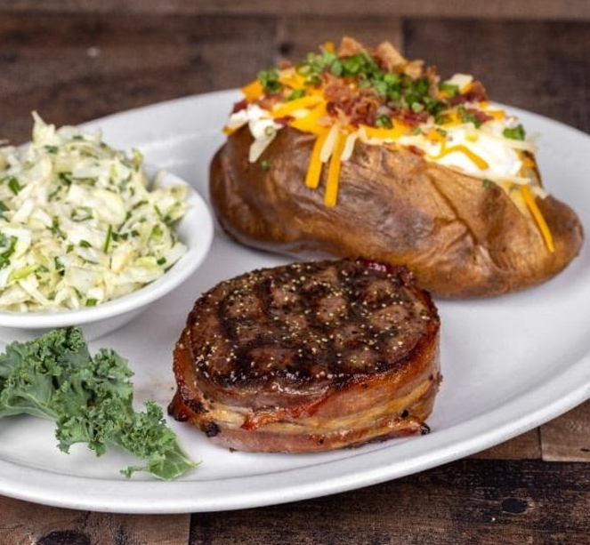 A grilled bacon-wrapped steak sits on a plate at a steakhouse on the West Coast, served with a loaded baked potato topped with cheese, sour cream, bacon, and chives, plus a bowl of coleslaw and a garnish of kale.