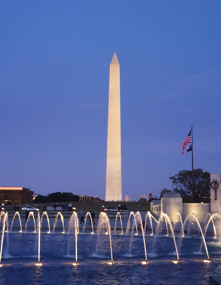 The Washington Monument at dusk, illuminated against a blue sky, stands as one of the top tourist attractions, with fountains and an American flag in the foreground.