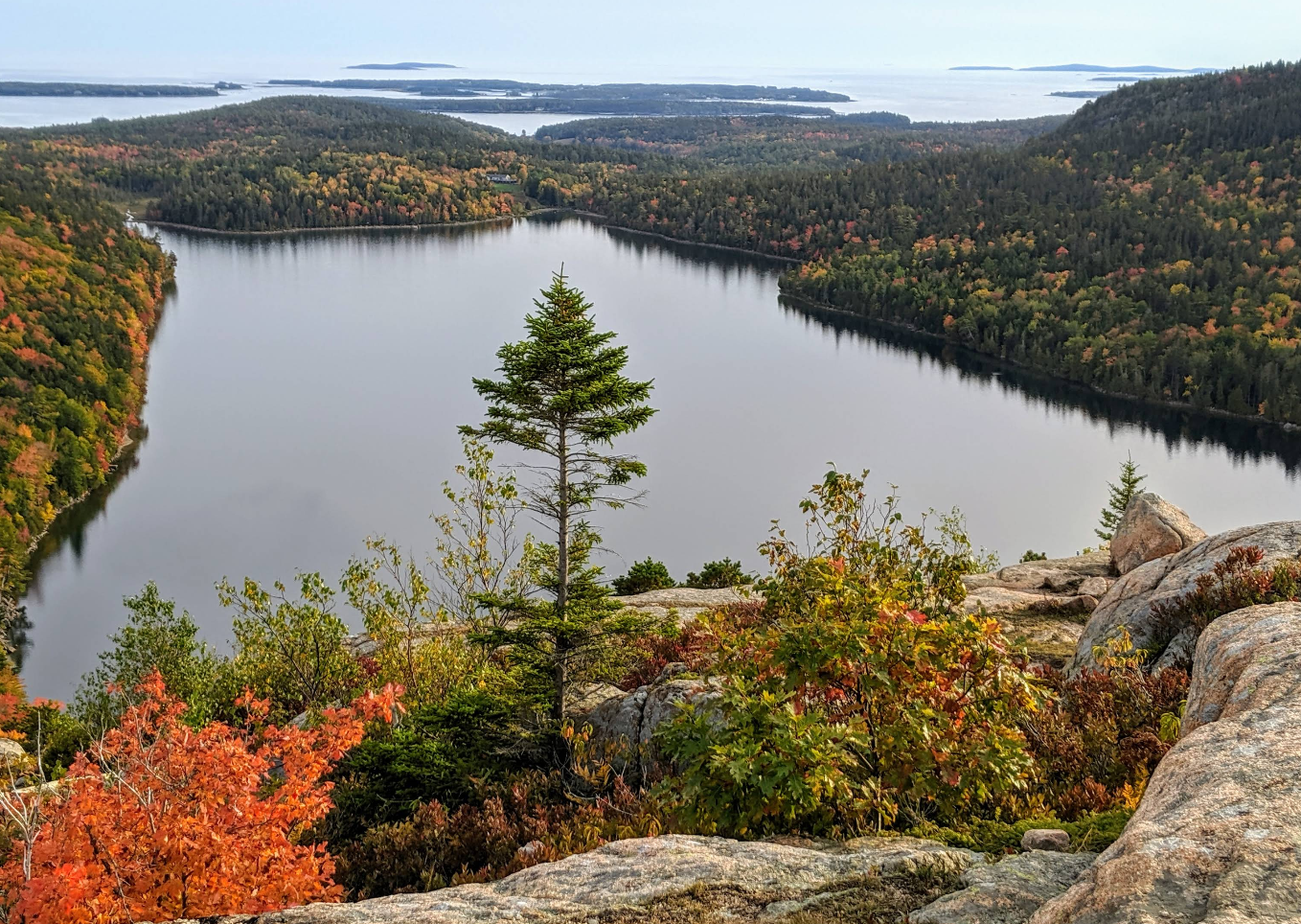 View from a rocky peak overlooking Jordan Pond in Acadia National Park during autumn, one of Maine's top tourist attractions. Vibrant fall foliage in reds and oranges surrounds the still water, dotted with distant islands.