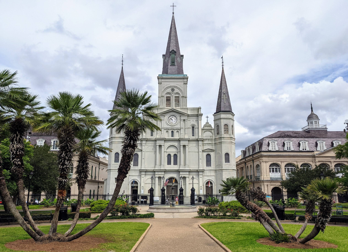 St. Louis Cathedral with three spires stands behind palm trees and a walkway in Jackson Square, one of New Orleans' top tourist attractions. The historic buildings flank the cathedral on both sides under a cloudy sky.