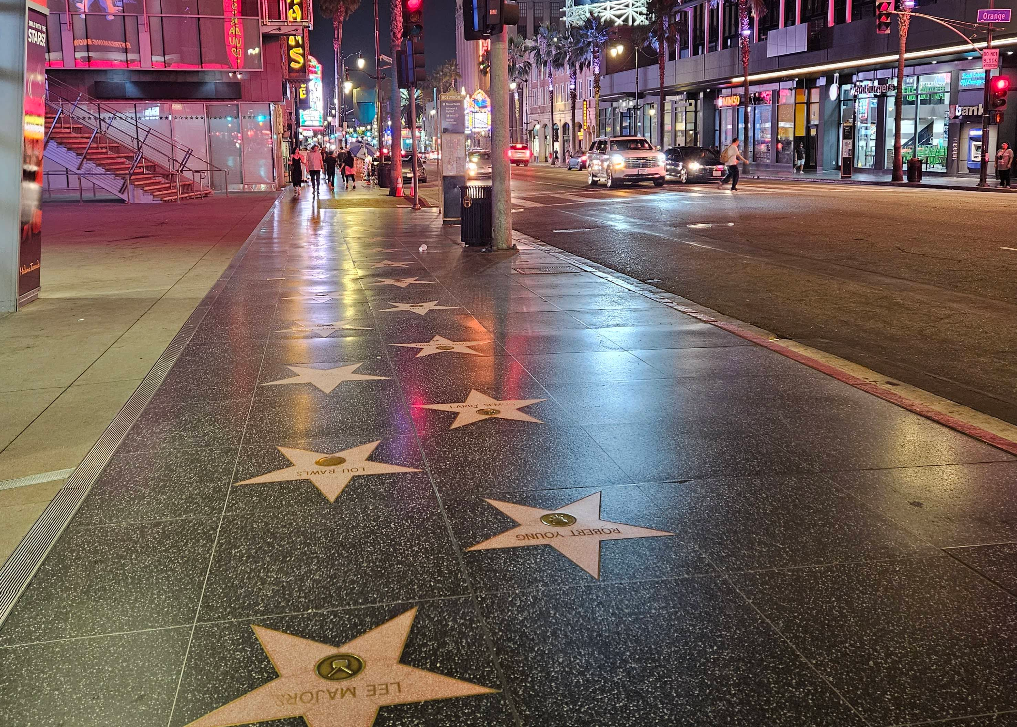 A nighttime street view of the Hollywood Walk of Fame, one of L.A.’s iconic tourist attractions, shows illuminated stars on the sidewalk. Cars and pedestrians pass by bright storefronts and glowing streetlights lining the bustling scene.