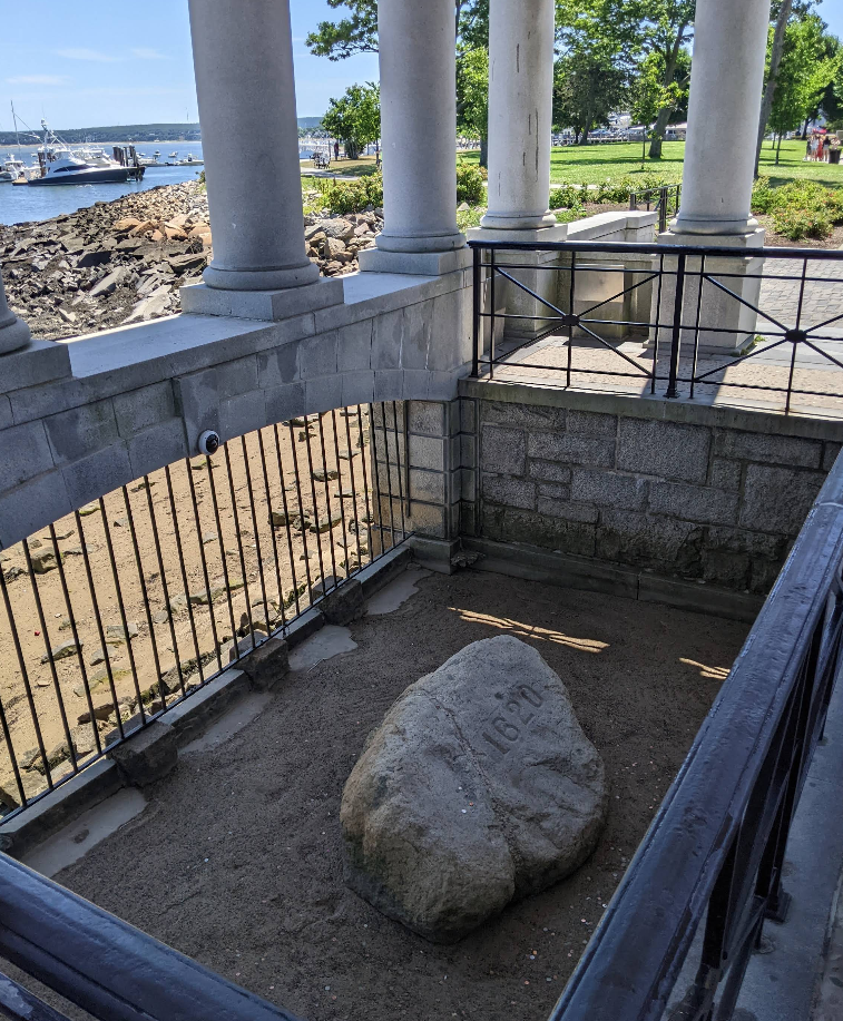 A large rock engraved with "1620" sits enclosed by a black metal fence and stone pillars, overlooking a waterfront area with trees, grass, and boats—one of the region’s most iconic tourist attractions.