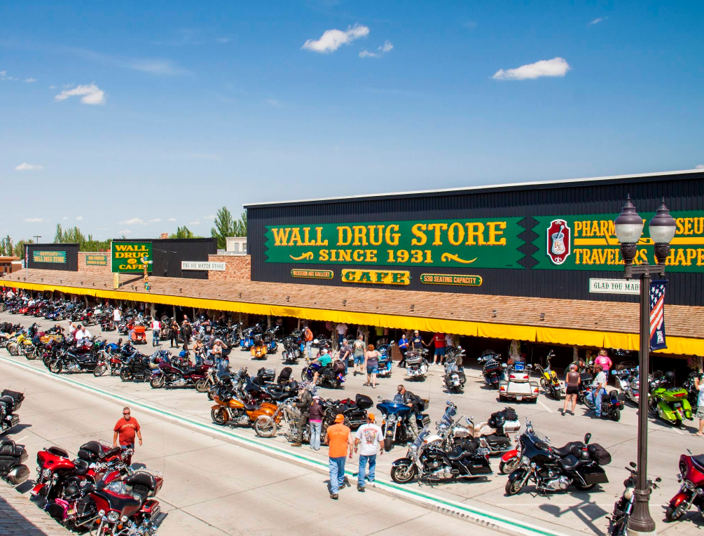 A busy street outside Wall Drug Store, one of the area's top tourist attractions, features rows of parked motorcycles and people walking along the sunny sidewalk. The store’s large sign is visible above the entrance.