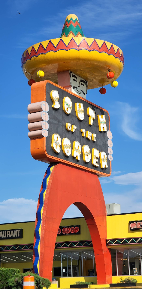 A large, colorful roadside sign shaped like a man in a sombrero holding a “South of the Border” sign stands as one of the iconic tourist attractions in front of yellow buildings under a clear blue sky.