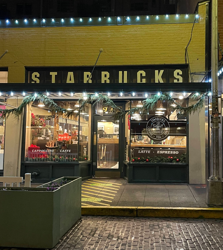 A warmly lit Starbucks storefront at night displays holiday decorations, greenery, and string lights—one of the cozy tourist attractions, with cobblestone pavement and a green planter in front. The sign above the entrance reads “STARBUCKS.”