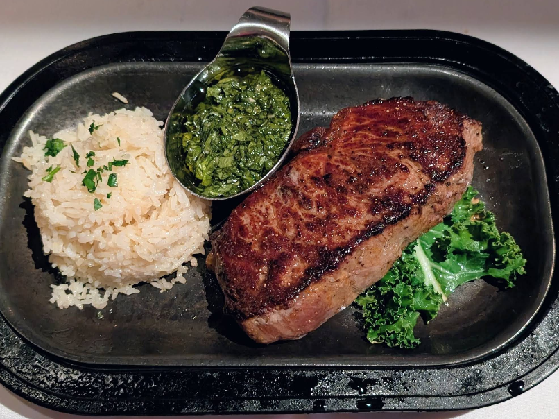 A plate from a steakhouse on the west coast featuring grilled steak, white rice topped with chopped herbs, leafy green garnish, and a metal sauce cup filled with vibrant green chimichurri sauce.