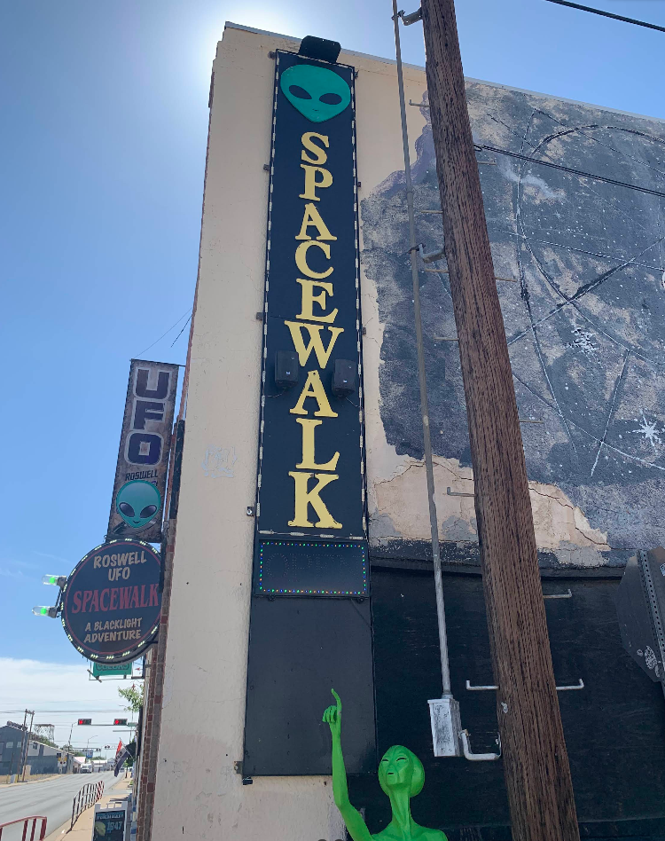 A tall black sign with a green alien face and yellow letters reading “SPACEWALK” is mounted on a building—one of the area’s quirky tourist attractions. Below, a green alien statue points upward, and a round “UFO Spacewalk” sign hangs nearby on the street.
