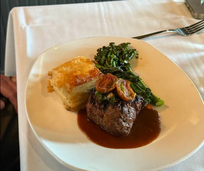 A plated meal from a steakhouse on the west coast features steak with sauce and mushrooms, layered potato gratin, and sautéed broccolini, all served on a white tablecloth with a fork nearby.