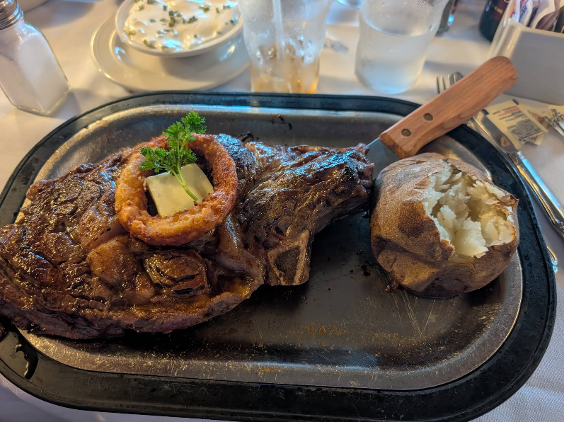 A cooked steak topped with butter, an onion ring, and parsley sits on a metal tray next to a baked potato—classic fare at a steakhouse on the West Coast. In the background are drinks, sour cream with chives, and cutlery on a white tablecloth.