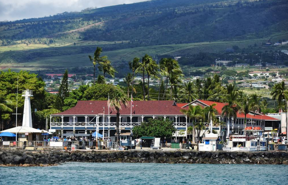 A waterfront building with a red roof and white trim sits among palm trees, near a rocky shoreline, with lush green hills rising in the background under a partly cloudy sky.