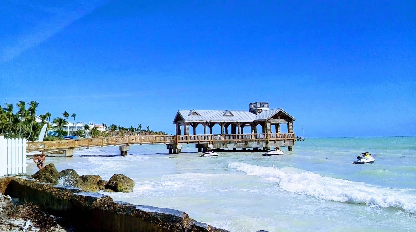 A wooden pier with a covered pavilion extends over turquoise ocean water, with jet skis riding nearby and palm trees lining the sandy shore under a clear blue sky.