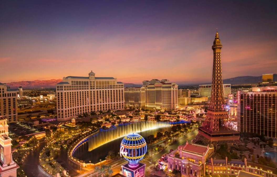 A vibrant view of the Las Vegas Strip at sunset, featuring illuminated hotels and casinos, the Bellagio fountains, and a replica of the Eiffel Tower against a colorful sky.