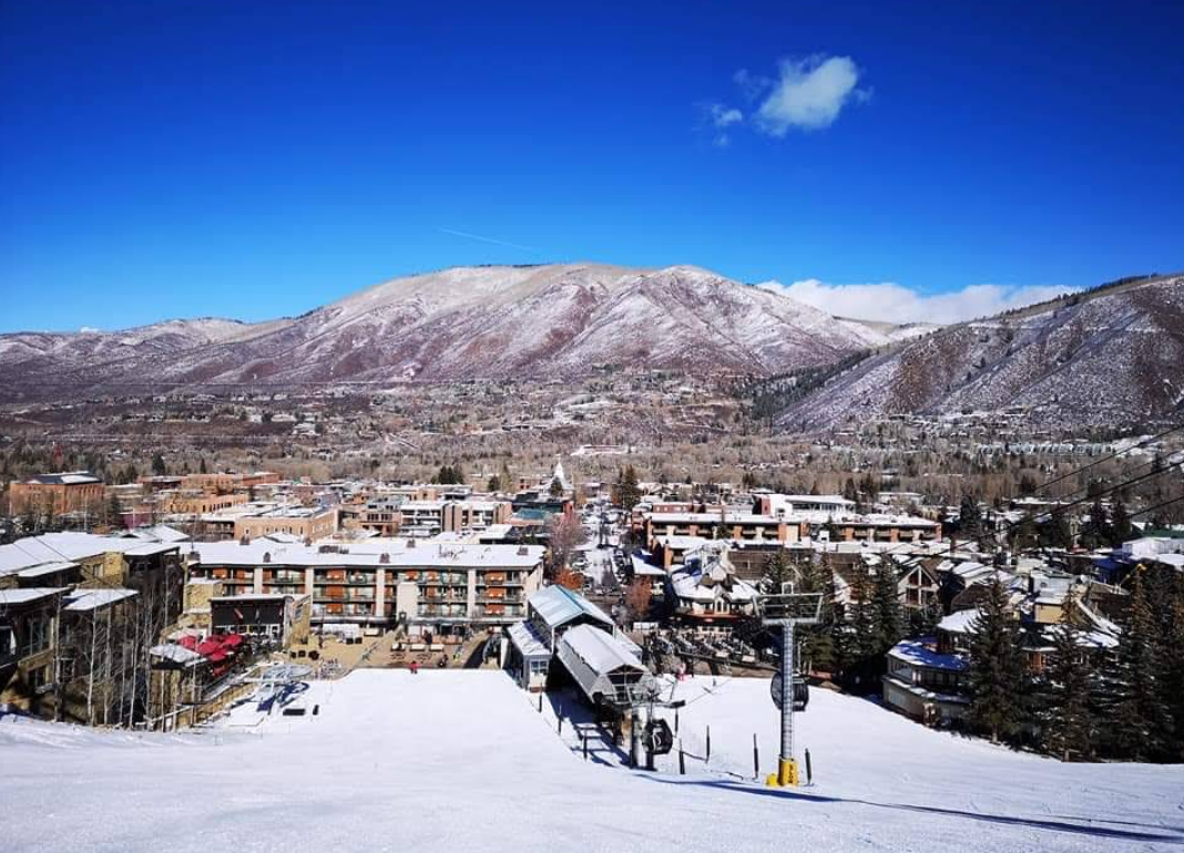 A snow-covered ski slope leads down to a small town with buildings, surrounded by leafless trees and hills. A ski lift is visible, with mountains and a bright blue sky in the background.