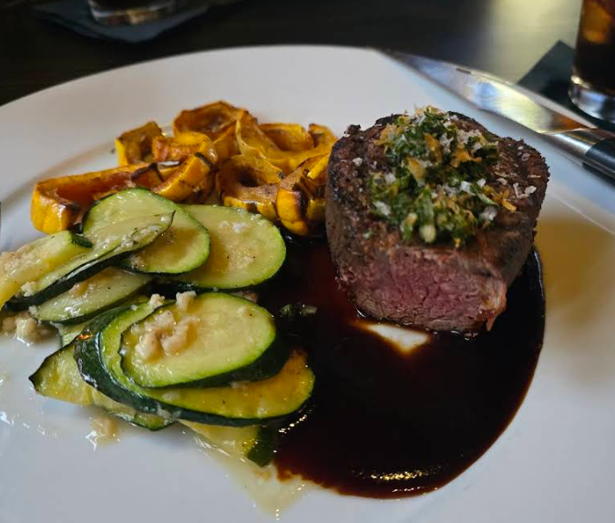 A plate with a medium-cooked steak topped with herbs and sauce, roasted squash, and sautéed zucchini, served on a white plate with a knife beside it—just like you'd find at a top steakhouse on the West Coast.
