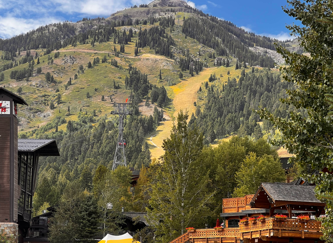 A scenic mountain covered with green trees and patches of yellow grass, with a ski lift tower visible. Wooden buildings and decks with flower boxes are in the foreground, and the sky is partly cloudy.