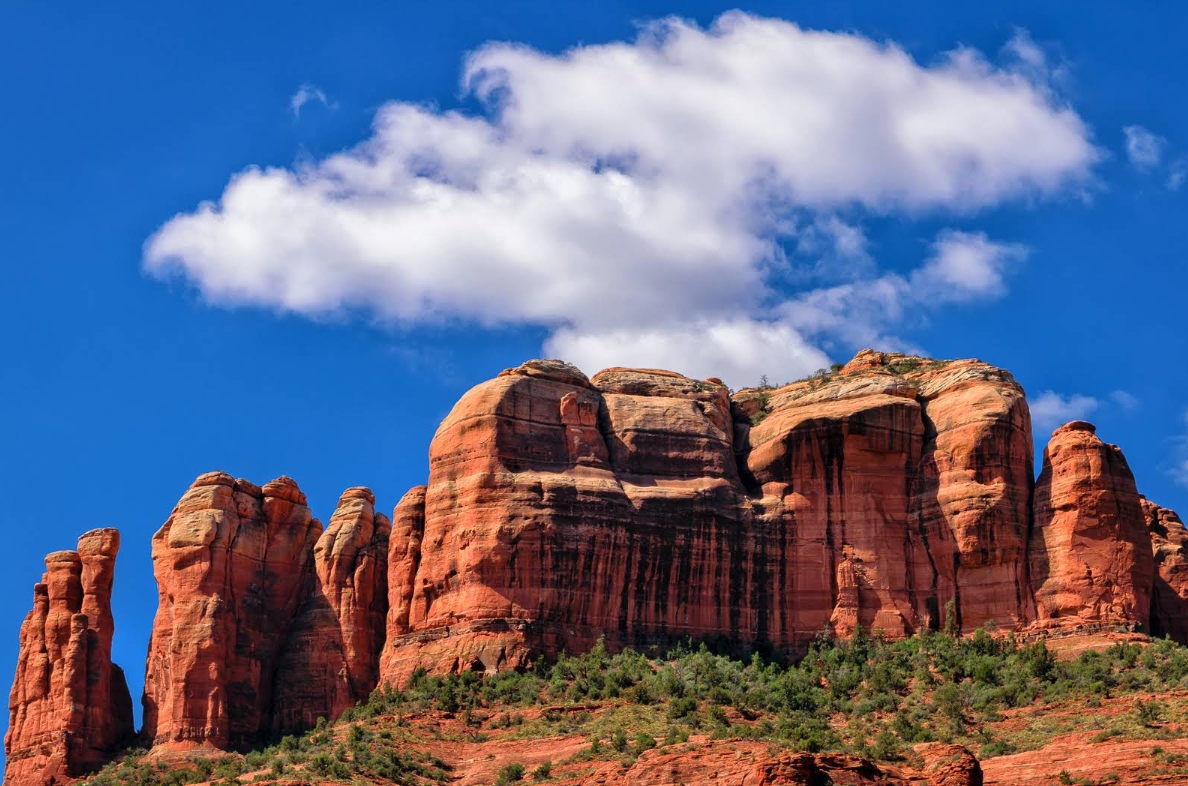 Red rock formations rise against a bright blue sky with a single white cloud above. Green vegetation grows at the base of the rocks, highlighting the vivid natural landscape.