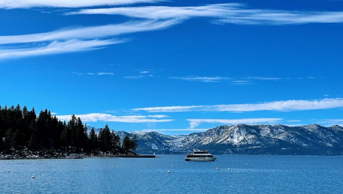A boat floats on a calm blue lake with snow-capped mountains and pine trees in the background under a bright blue sky with wispy clouds.