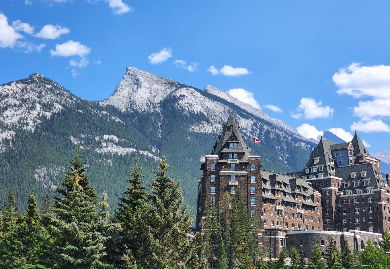 A large historic hotel with pointed roofs stands among pine trees, with rugged, snow-dusted mountains and a blue sky with scattered clouds in the background. A Canadian flag is visible on the building.