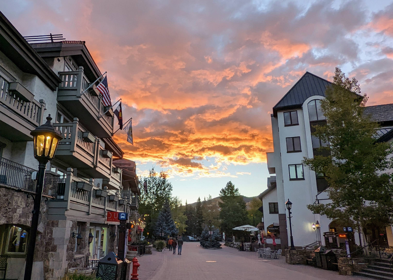 A pedestrian street in a mountain town at sunset, with vibrant orange clouds in the sky, flanked by buildings with balconies and flags, trees, and a few people walking.