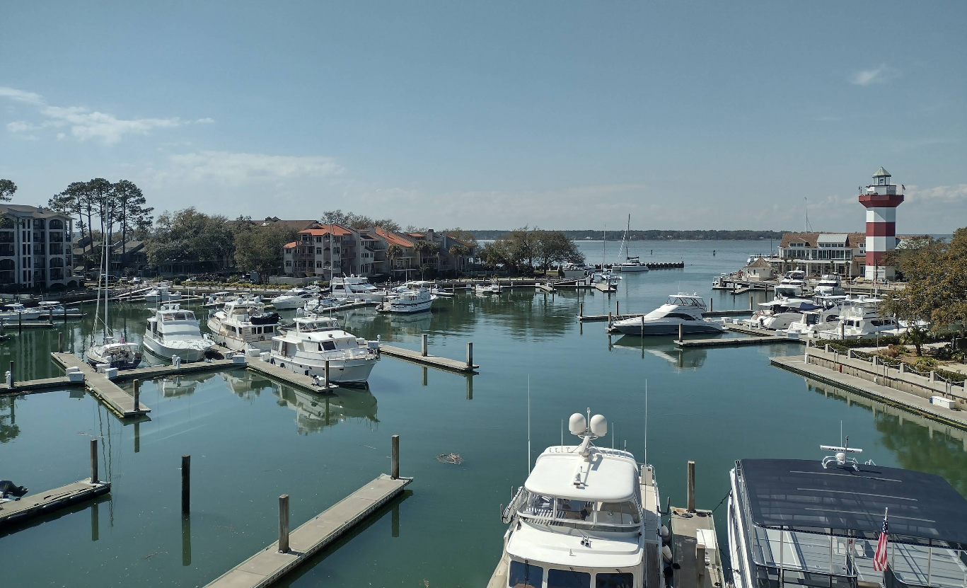 A marina with several docked boats and yachts, calm water, waterfront buildings, and a red-and-white striped lighthouse on the right under a partly cloudy sky.